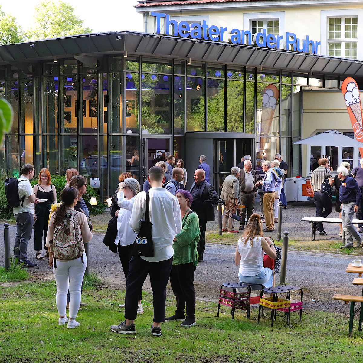 Foto von der Abschlussfeier des Festivals "Stuecke" vor dem Theater an der Ruhr. Man sieht Menschen, die sich in kleinen Gruppen mit einem Glas in der Hand unterhalten.